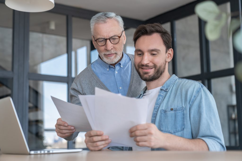 Caring loving adult son helping his old senior elderly father paying bills, counting money, doing paperwork at home together. Aid to elderly generation. Happy father`s day!