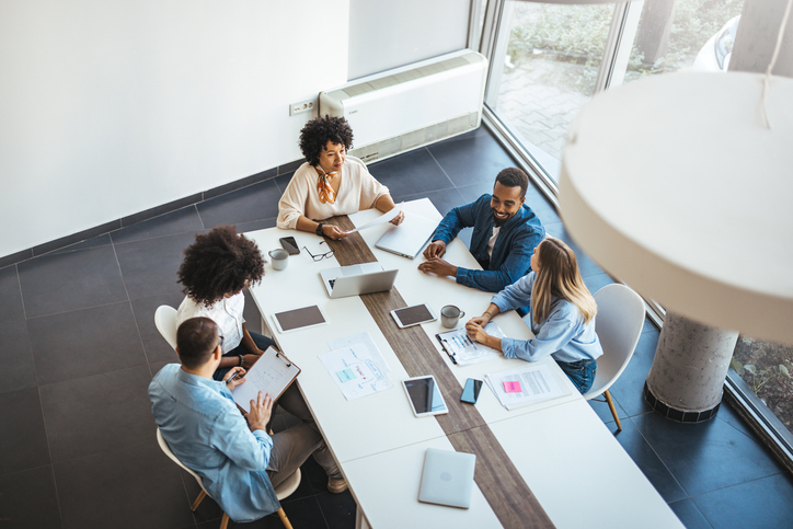 A diverse group of professionals engaged in a collaborative meeting in a modern office setting. They are discussing ideas with devices and documents on a table, showcasing teamwork and interaction.